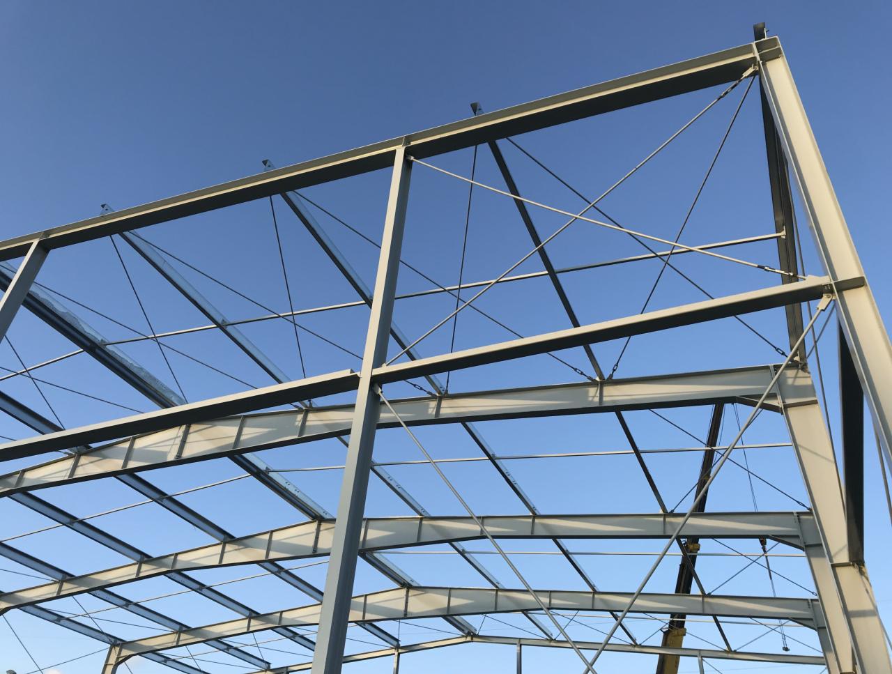 Szkielet powstającej hali z belek stalowych, widok z dołu na błękitne niebo, widoczne elementy mocujące i liny dźwigu.