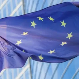 Flaga Unii Europejskiej powiewająca na tle nowoczesnego budynku biurowego z przeszkloną fasadą. Widoczne maszt i fragment błękitnego nieba.