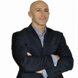 Mężczyzna łysy w ciemnej marynarce i koszuli w kratę, z założonymi rękami na białym tle, uśmiecha się delikatnie. Portret biznesowy.