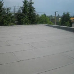 Szary, płaski dach pokryty papą termozgrzewalną z widocznym narzędziem pracy, na tle zieleni drzew i zabudowań.