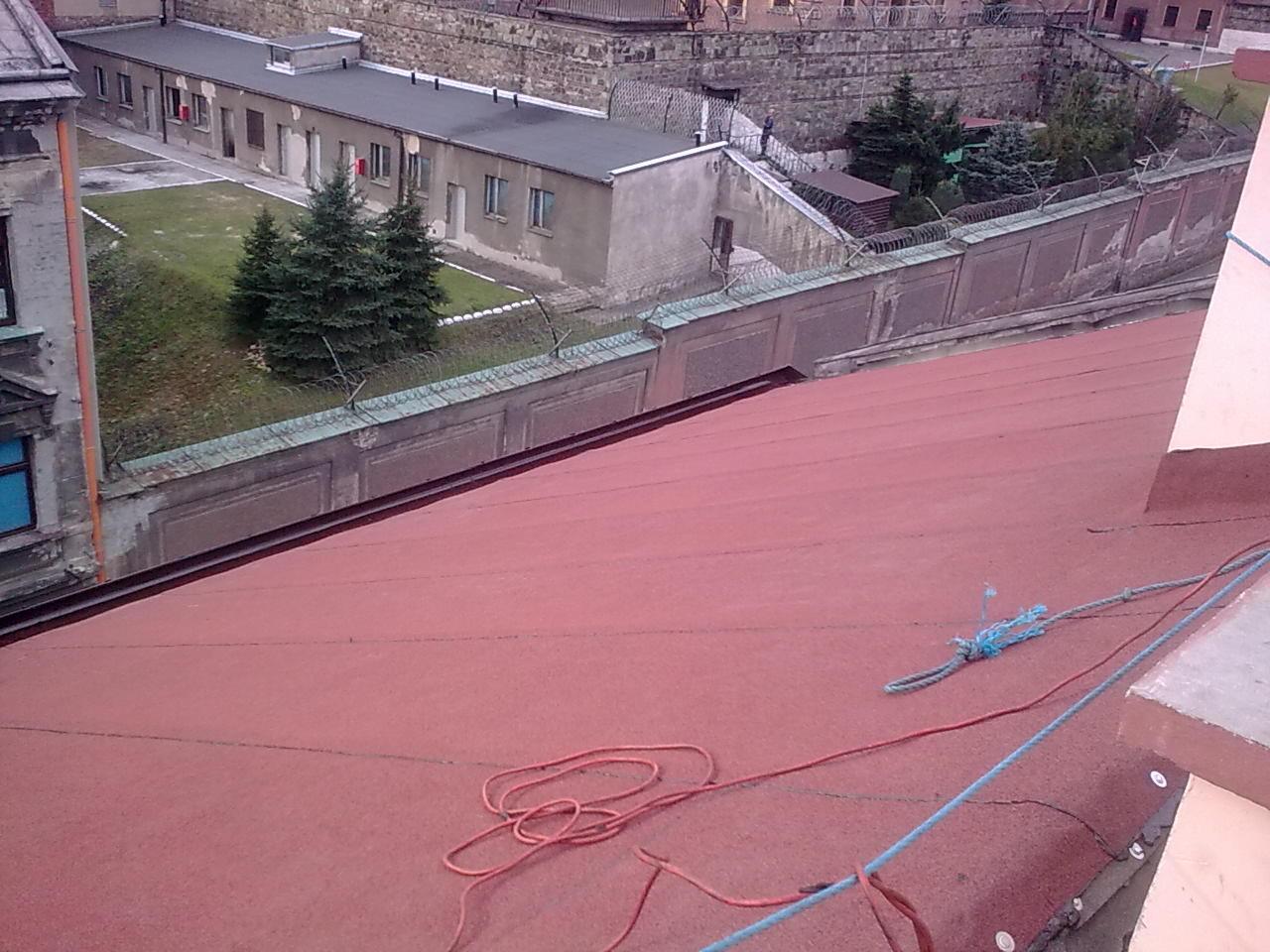 Czerwony dach budynku z widocznymi elementami zabezpieczeń i otoczeniem z budynkami i zielenią. Widok z góry ukazuje strukturę pokrycia.