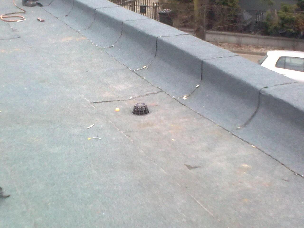 Płaski, pokryty papą dach z widocznym odpływem wody. Elementy wykończeniowe dachu wzdłuż krawędzi. Widoczne drobne zabrudzenia i pozostałości po pracy.