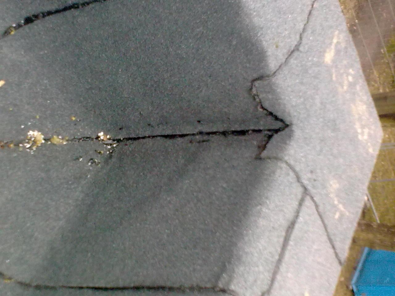 Zbliżenie na fragment dachu pokrytego papą z widocznymi pęknięciami i uszkodzeniami, sugerującymi potrzebę naprawy lub wymiany, z lekkim widokiem na otoczenie.