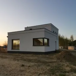 Nowoczesny, biały dom jednorodzinny z płaskim dachem, widok z zewnątrz na tle piaszczystego terenu w porannym świetle, z widocznymi oknami z ciemnymi ramami i minimalistyczną architekturą.