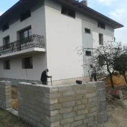 Dwukondygnacyjny dom z szarą elewacją i balkonem z ozdobną balustradą; w tle drzewo bez liści i pracownicy budowlani przy wznoszeniu muru z bloczków betonowych, w pobliżu leży czerwona łopata