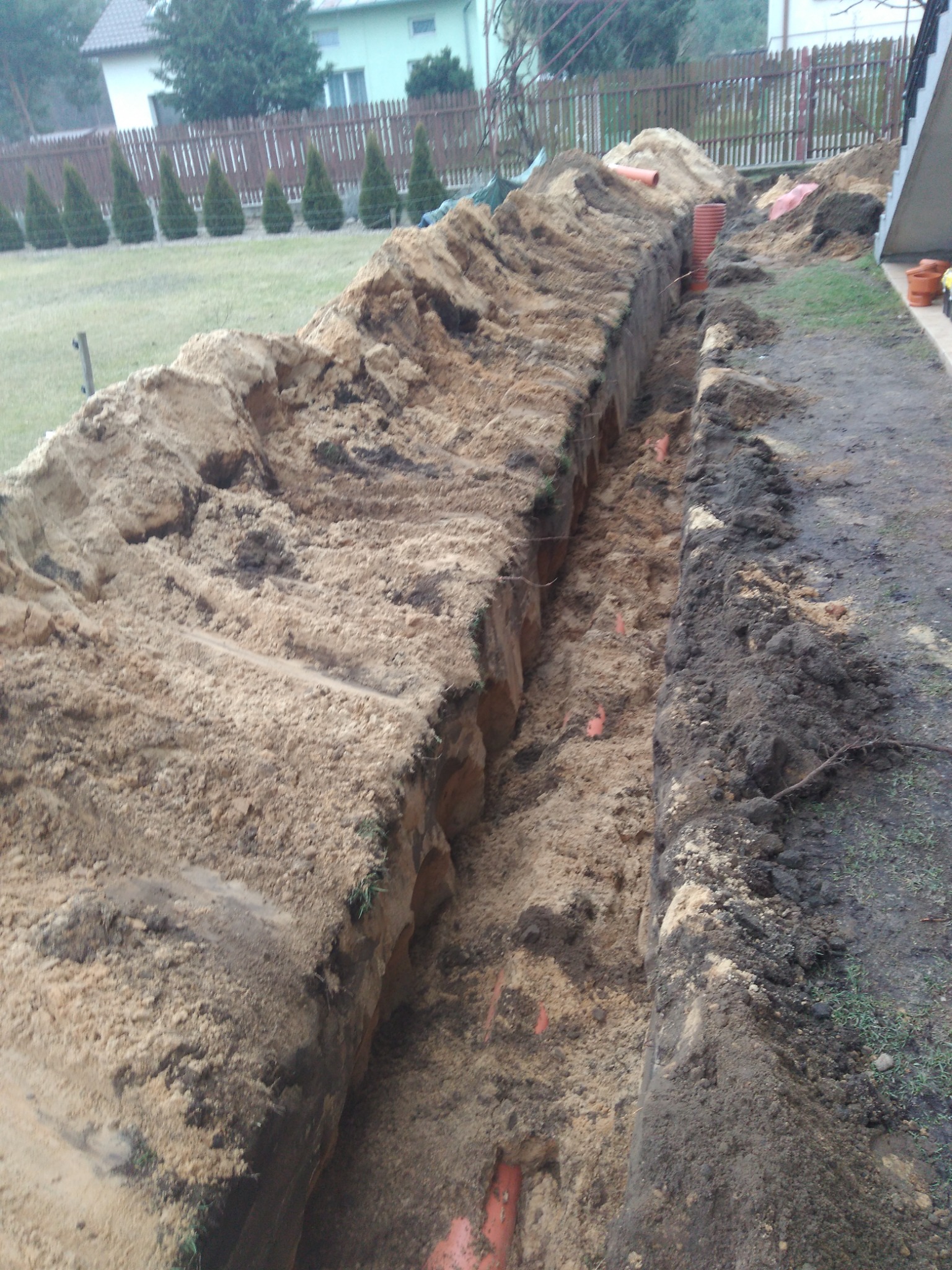 Długi wykop w ziemi z widocznymi pomarańczowymi rurami instalacyjnymi na dnie, obok trawnik z tujami i ogrodzeniem.