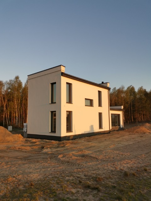 Nowoczesny, dwukondygnacyjny dom z płaskim dachem, pomalowany na biało, z ciemnymi ramami okiennymi, sfotografowany o zachodzie słońca na piaszczystej działce z widocznym lasem w tle.