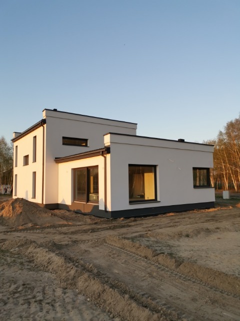 Nowoczesny, dwukondygnacyjny dom z płaskim dachem, pomalowany na biało, z dużymi oknami z czarnymi ramami, widoczny w porannym słońcu na działce z piaskiem.