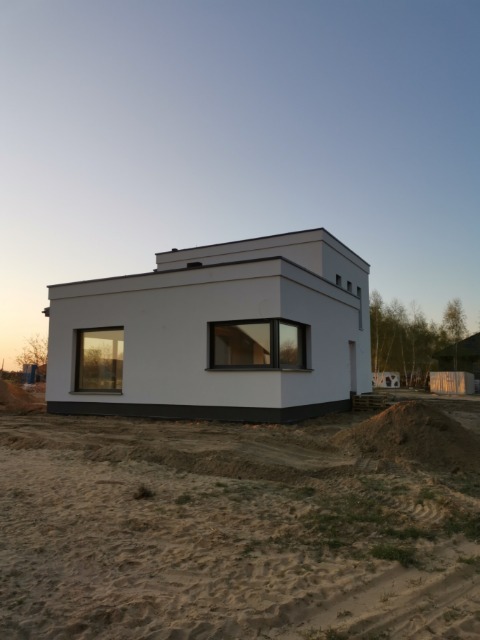 Nowoczesny, biały dom jednorodzinny z płaskim dachem, widok z zewnątrz na tle piaszczystego terenu w porannym świetle, z widocznymi oknami z ciemnymi ramami i minimalistyczną architekturą.