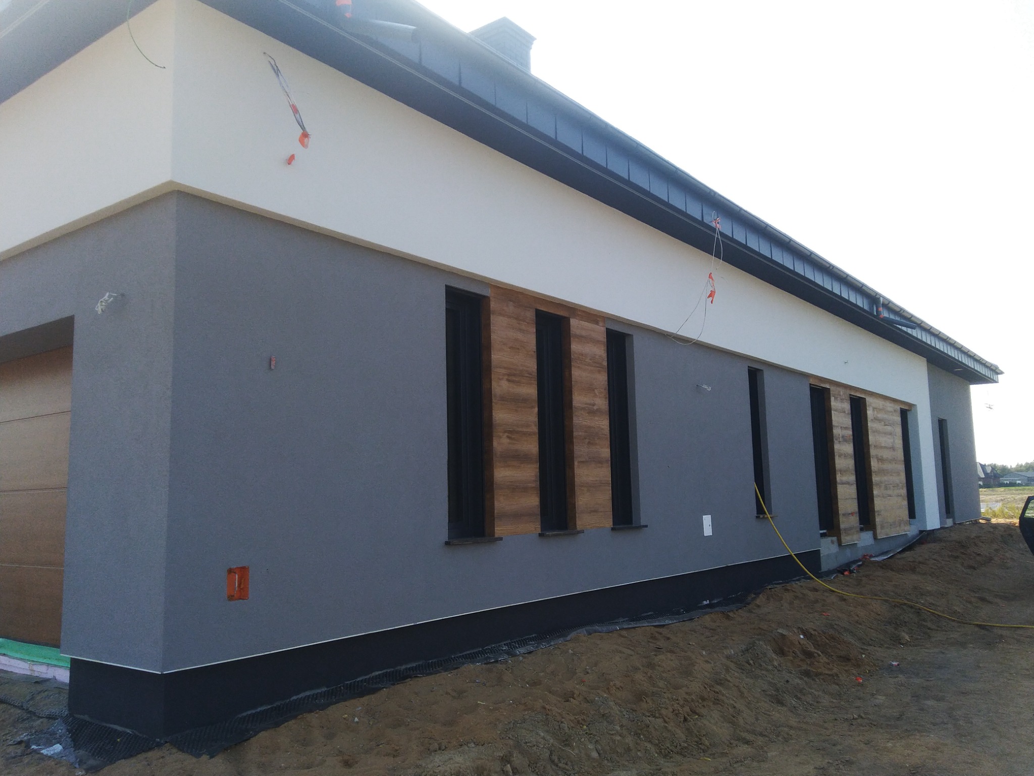 Nowoczesny dom jednorodzinny z szarą elewacją i drewnianymi panelami dekoracyjnymi wokół okien, widoczne przygotowania terenu wokół budynku, w tym drenaż przy fundamentach.