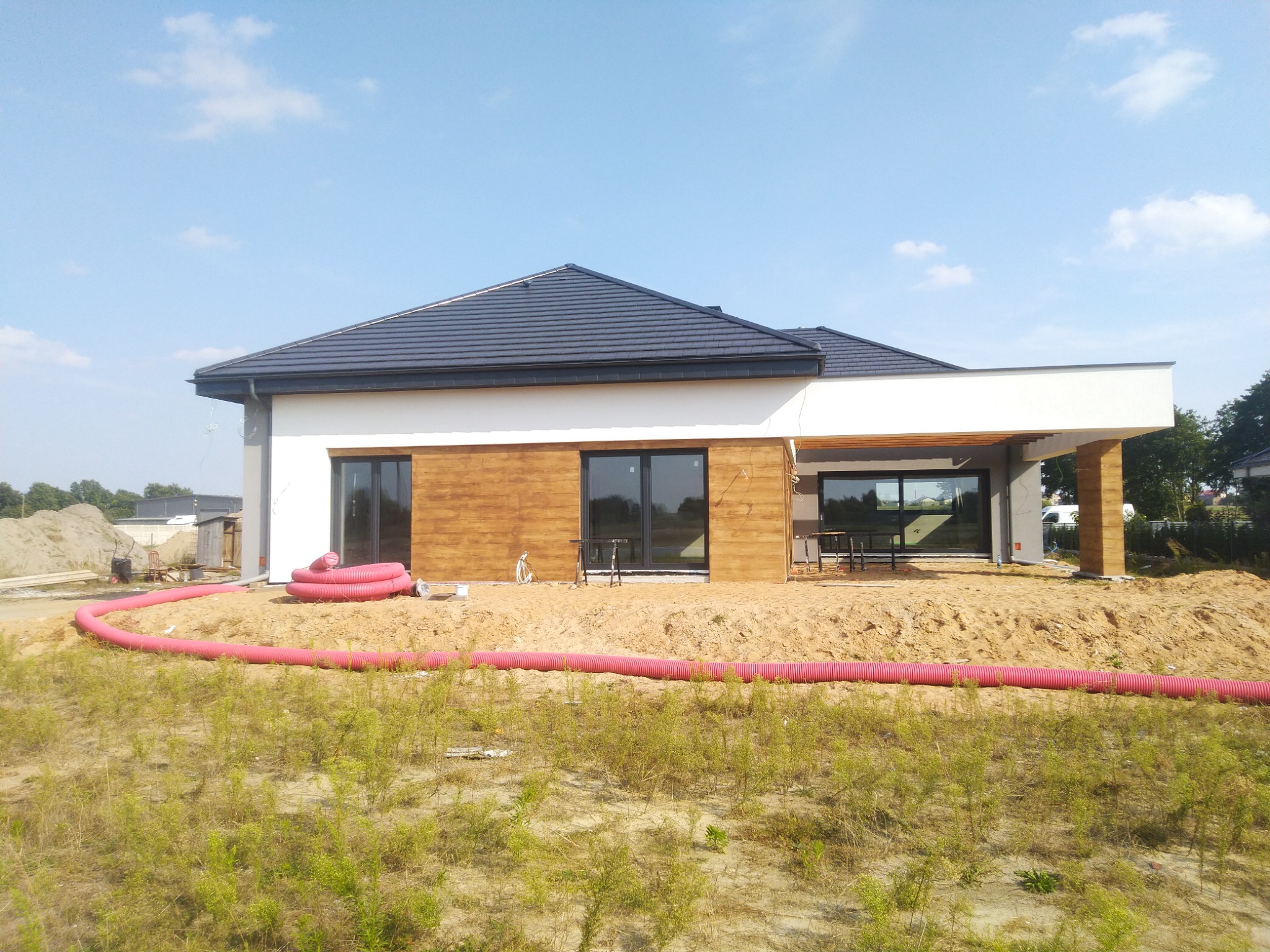 Nowoczesny dom jednorodzinny w trakcie budowy, z widocznymi elementami elewacji drewnianej, dużymi oknami i czerwonymi rurami instalacyjnymi na ziemi.