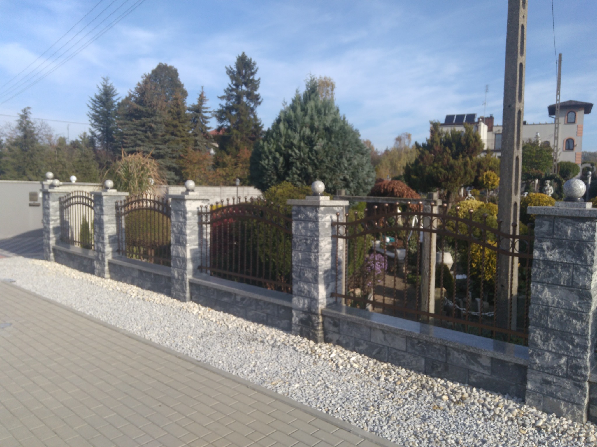 Nowoczesne ogrodzenie z szarej kostki i metalowymi przęsłami na tle zadbanego ogrodu z domem w oddali.