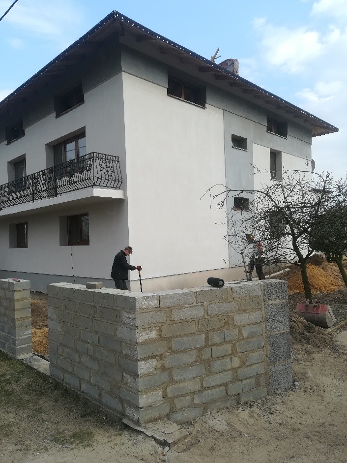 Dwukondygnacyjny dom z szarą elewacją i balkonem z ozdobną balustradą; w tle drzewo bez liści i pracownicy budowlani przy wznoszeniu muru z bloczków betonowych, w pobliżu leży czerwona łopata