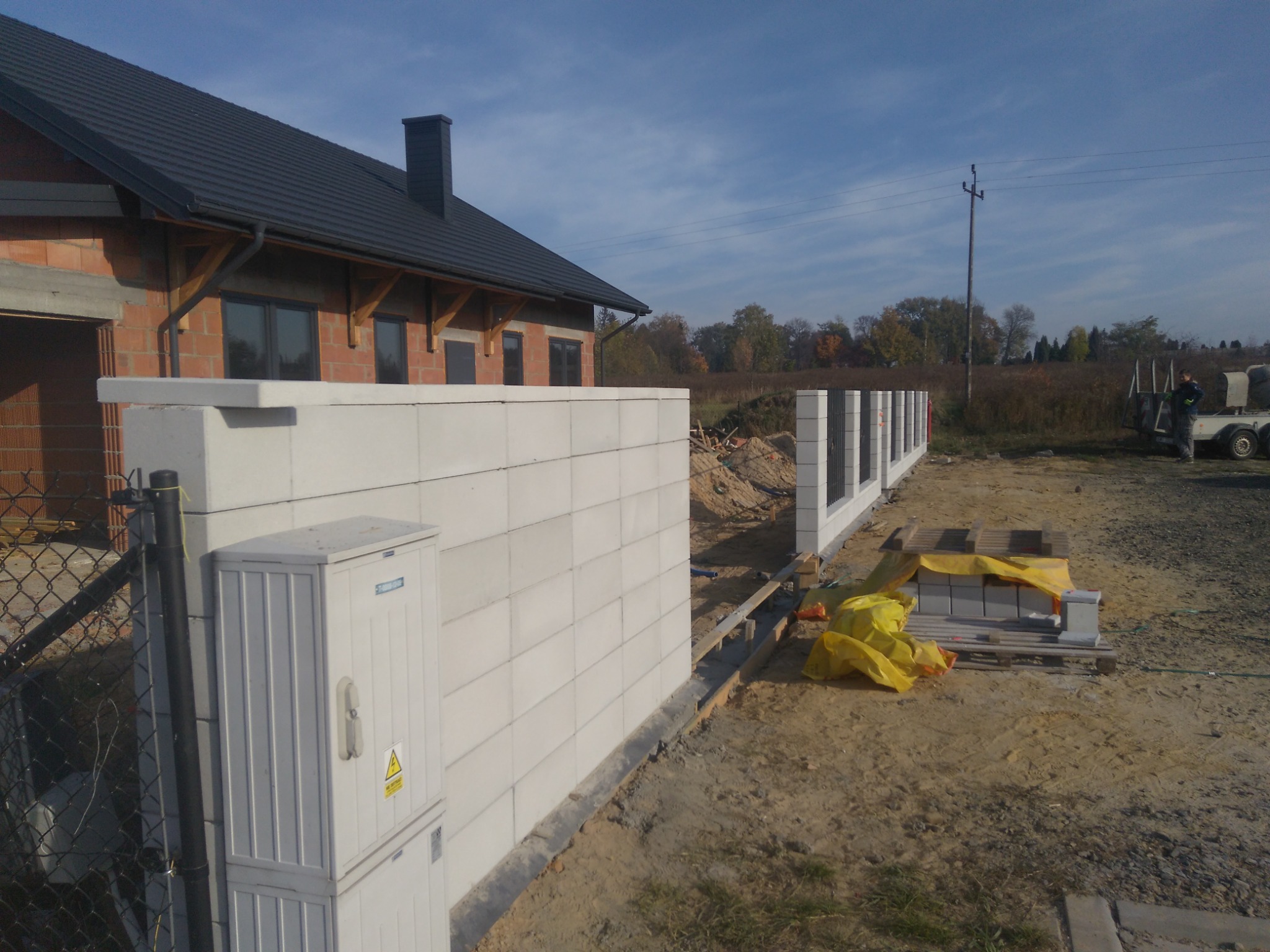 Wznoszenie muru z bloczków betonowych przy nowo budowanym domu z cegły, widoczna skrzynka elektryczna, paleta z bloczkami zabezpieczona żółtą folią, w tle słup elektryczny i częściowo zalesiony teren.