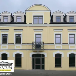 Żółta, odnowiona elewacja budynku z dekoracyjnymi elementami architektonicznymi i balkonem. Widoczne okna i mansardy. Budynek po renowacji.