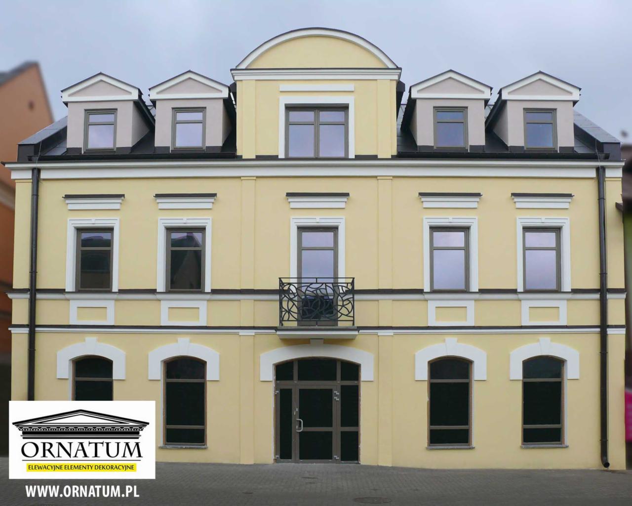 Żółta, odnowiona elewacja budynku z dekoracyjnymi elementami architektonicznymi i balkonem. Widoczne okna i mansardy. Budynek po renowacji.