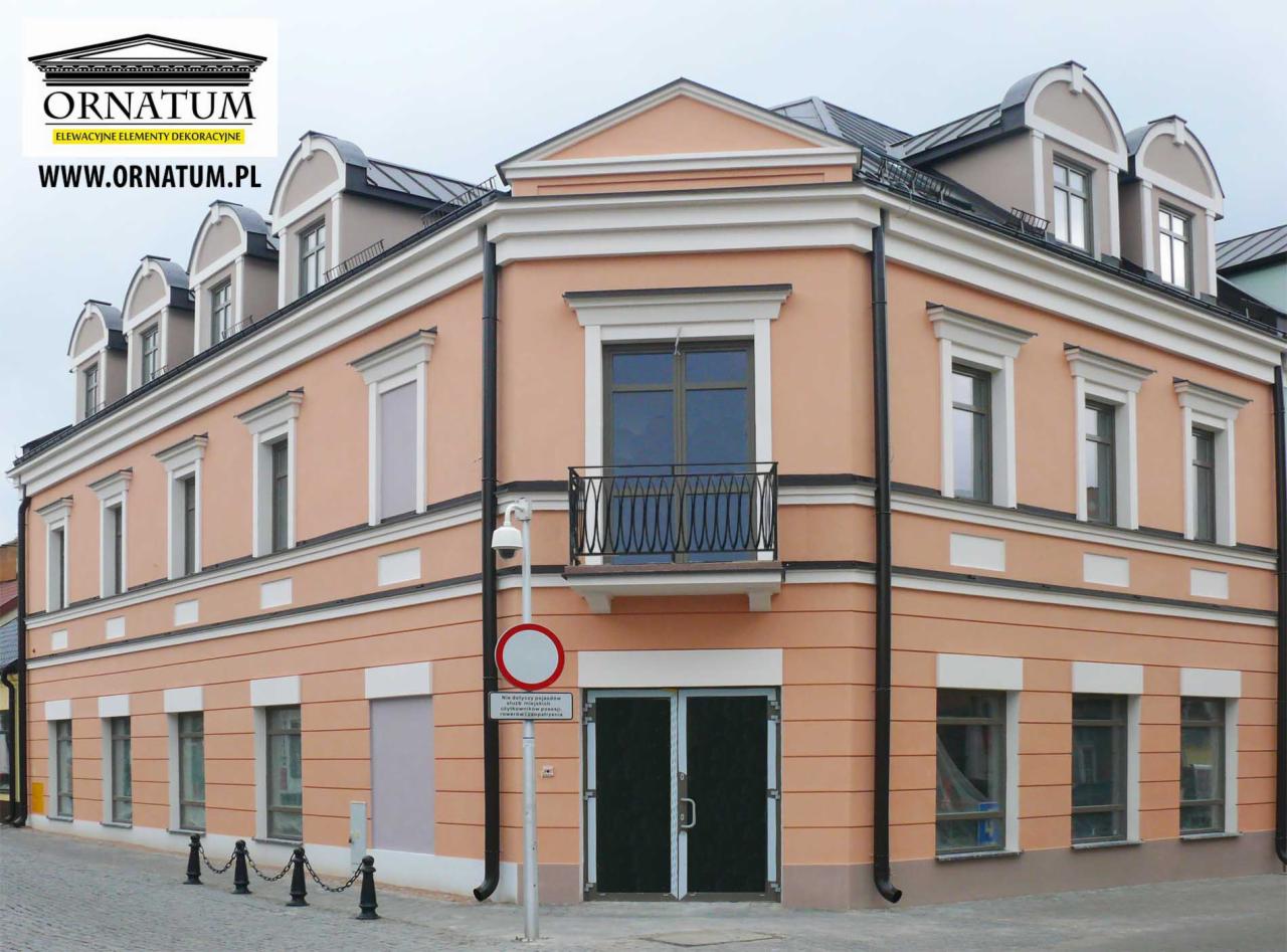 Odnowiona elewacja budynku w kolorze brzoskwini z białymi detalami architektonicznymi, balkonem i czarną stolarką okienną. Widoczny znak drogowy.