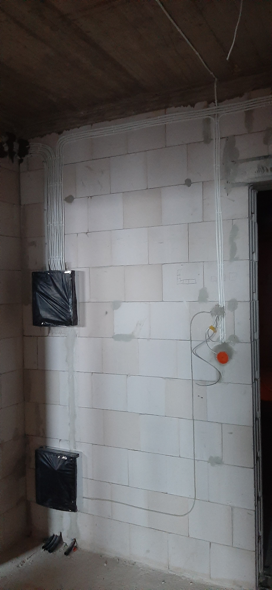 Świeżo położona instalacja elektryczna w surowym wnętrzu z białych bloczków, kable w peszlach prowadzone po ścianach i suficie, puszki elektryczne zabezpieczone czarną folią.