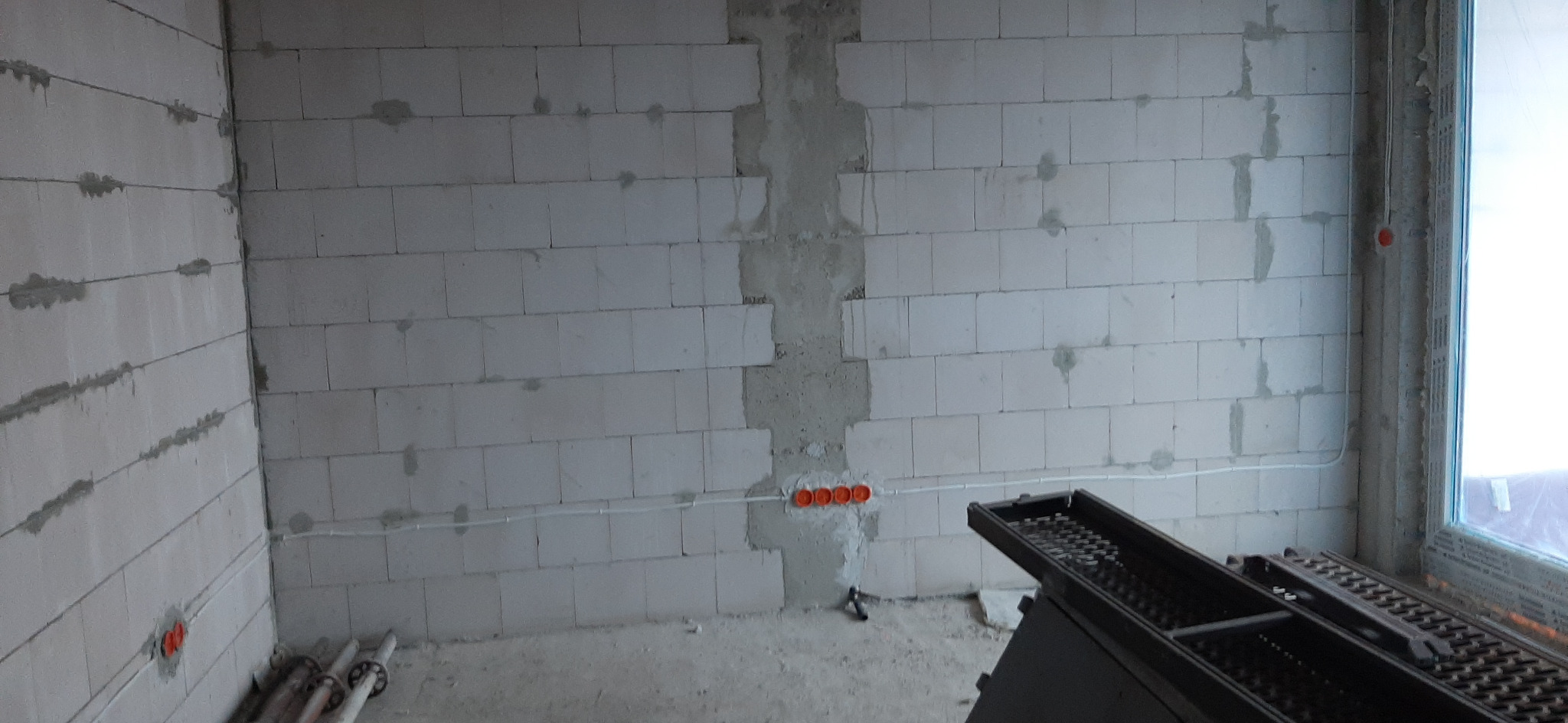 Surowy stan wnętrza z widoczną instalacją elektryczną: przewody w białej listwie przypodłogowej i pomarańczowe puszki podtynkowe w ścianie z bloczków betonowych.