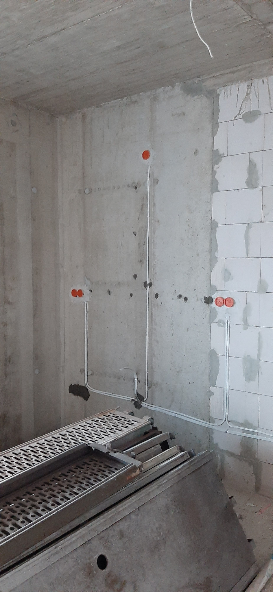 Surowy stan: Przewody elektryczne w białych rurkach prowadzone po surowej ścianie z betonu komórkowego, widoczne pomarańczowe puszki instalacyjne, na pierwszym planie metalowe podesty robocze.