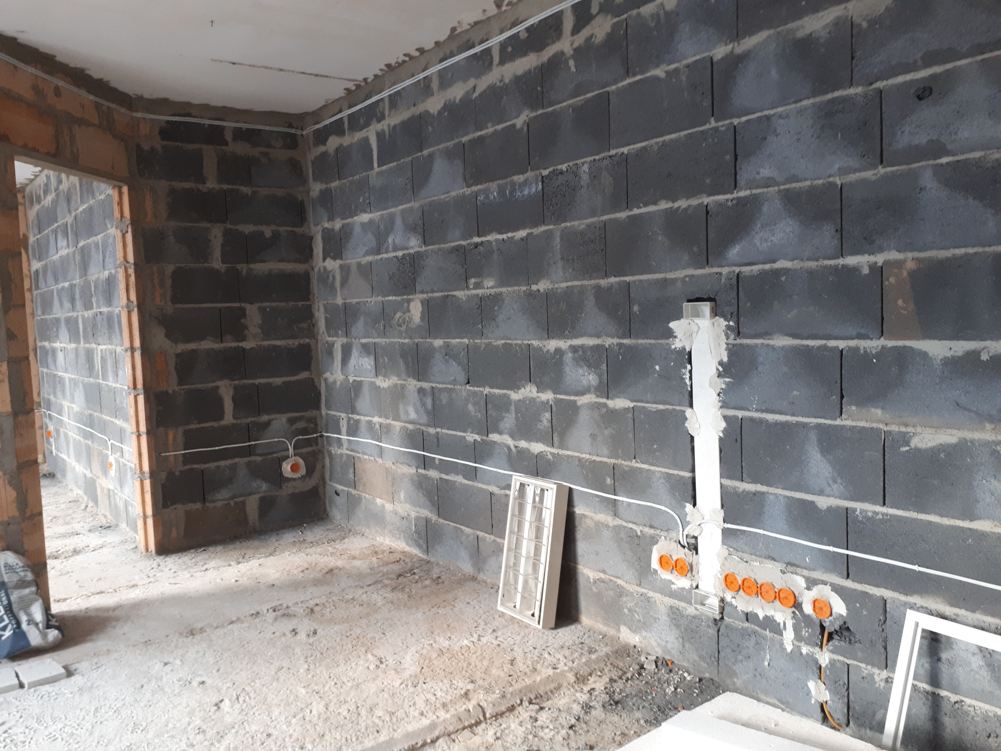 Surowa ściana z szarych bloczków z widoczną instalacją elektryczną: kable w peszlach, puszki podtynkowe z pomarańczowymi wkładkami, pionowy kanał kablowy zalany gipsem, w tle ościeżnica drzwiowa...