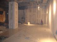 Surowe wnętrze w trakcie prac wykończeniowych: betonowa podłoga, oświetlone ściany, widoczne kable i kolumna. W oddali majster w czerwonej koszulce.