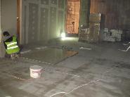 Pracownik w kamizelce odblaskowej przygotowuje podłoże pod posadzkę w surowym wnętrzu. Widoczne płyty, kable i wiadro z farbą. W tle materiały budowlane.