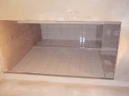 Wnęka wyłożona jasnymi, kwadratowymi płytkami ceramicznymi. Widoczne świeże fugi i delikatne odbicia światła na powierzchni płytek. Minimalistyczne wykończenie.