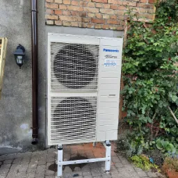 Zewnętrzna jednostka klimatyzacji Panasonic Inverter, zamontowana na białym stelażu przy ceglanej ścianie z pnącą roślinnością i betonowej ścianie z rynną spustową, na kostce brukowej.