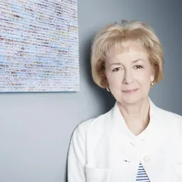 Portret kobiety w białym żakiecie, z obrazem w tle, w stonowanej kolorystyce. Kobieta uśmiecha się delikatnie, ma krótkie blond włosy i subtelny makijaż.