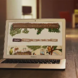 Laptop MacBook Air wyświetlający stronę internetową z ilustracjami i tekstem 'Muzyczny Elementarz dla Dzieci', umieszczony na drewnianym biurku.