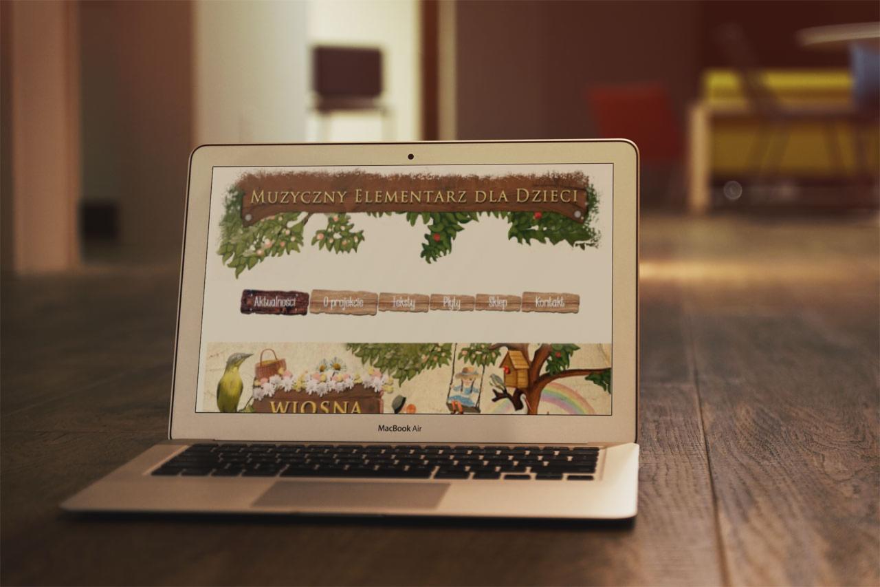 Laptop MacBook Air wyświetlający stronę internetową z ilustracjami i tekstem 'Muzyczny Elementarz dla Dzieci', umieszczony na drewnianym biurku.