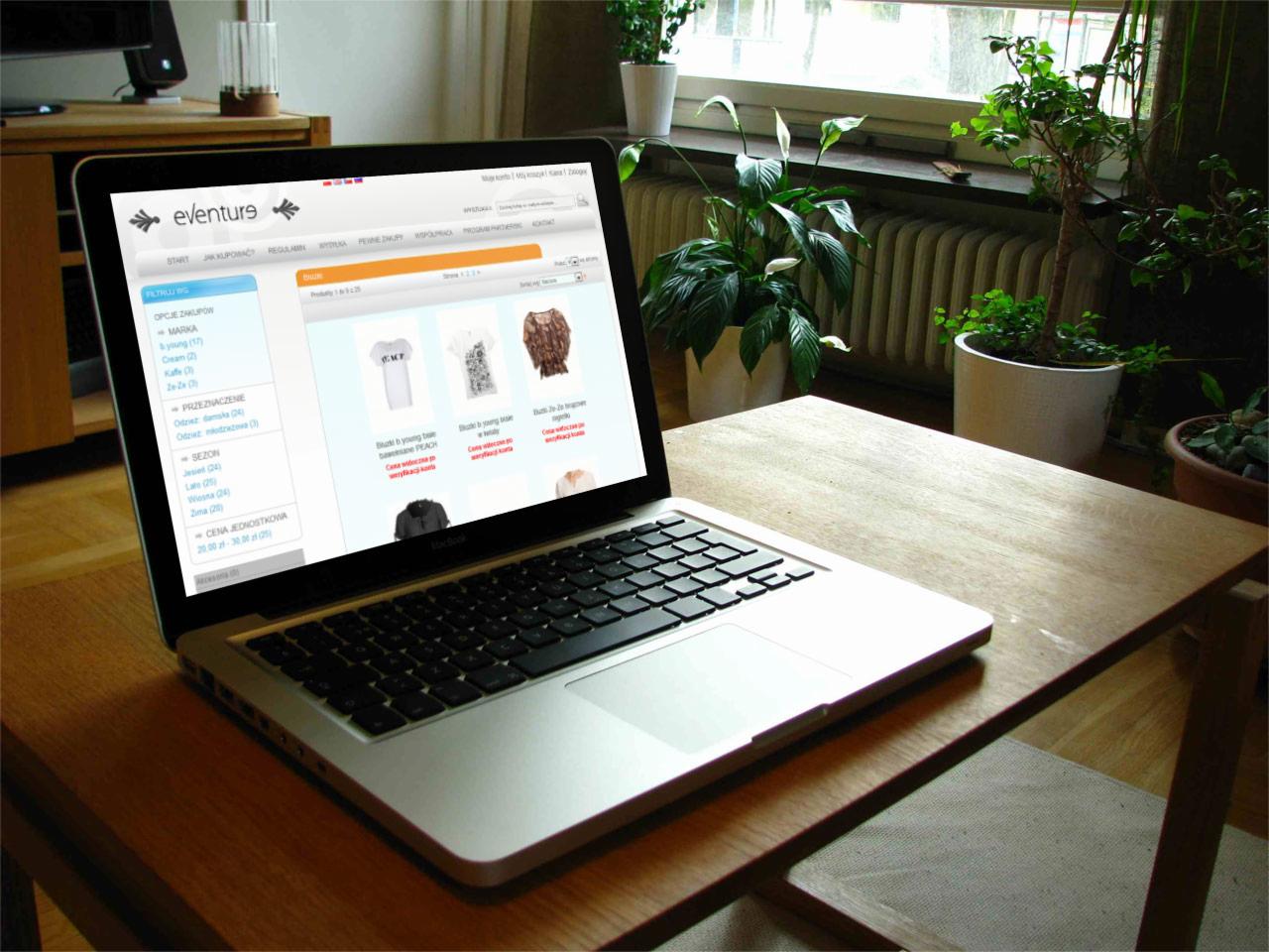 Laptop MacBook wyświetla stronę sklepu internetowego Eventure z ofertą odzieży, ustawiony na drewnianym biurku w pobliżu okna z roślinami doniczkowymi.
