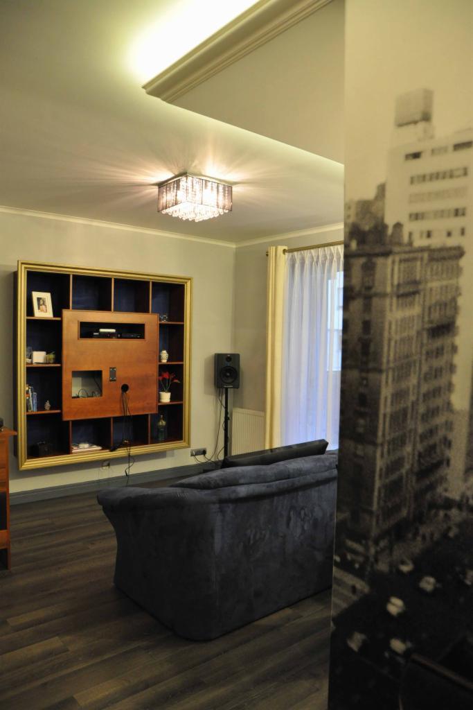 Salon z ciemną podłogą, szarą sofą i regałem w złotej ramie. Na ścianie tapeta z motywem miejskim. Centralnie żyrandol. Widok na okno z zasłoną.