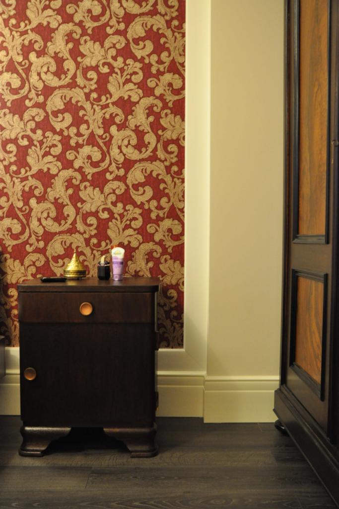 Ciemna szafka nocna z dekoracyjnym blatem, na tle wzorzystej tapety w odcieniach czerwieni i złota, obok fragmentu drewnianej szafy. Uwagę zwraca oświetlenie pomieszczenia.