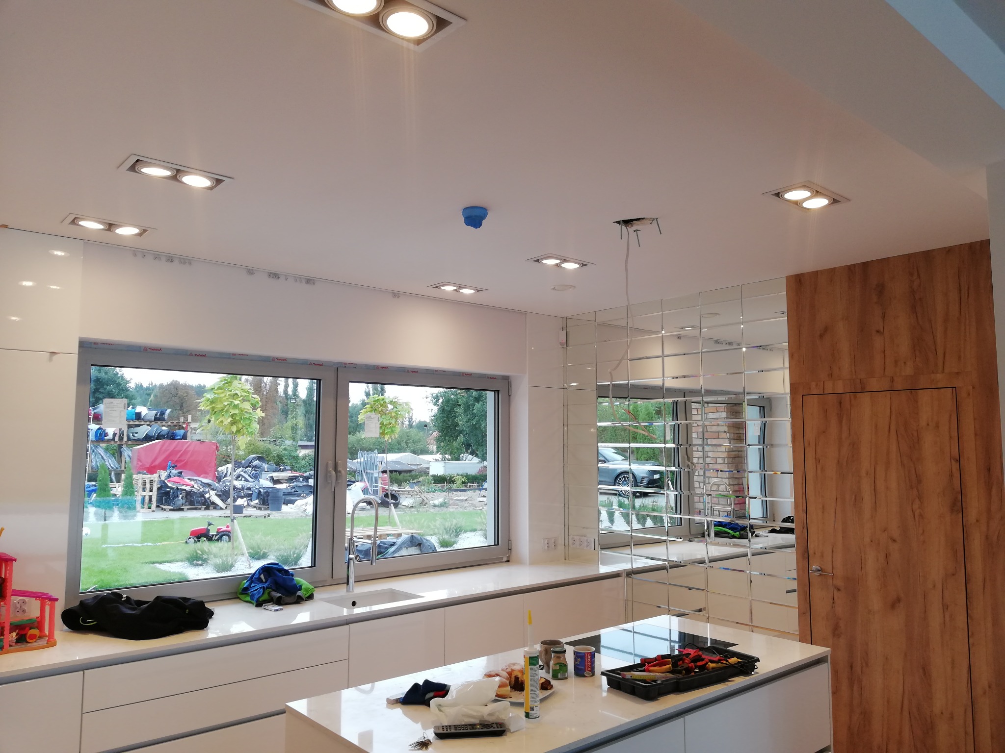 Nowoczesna kuchnia z białymi szafkami, dużym oknem z widokiem na podwórze z kosiarką, oraz zamontowanym oświetleniem sufitowym typu downlight, czujnik dymu oraz przygotowane miejsce na lampę wiszącą.