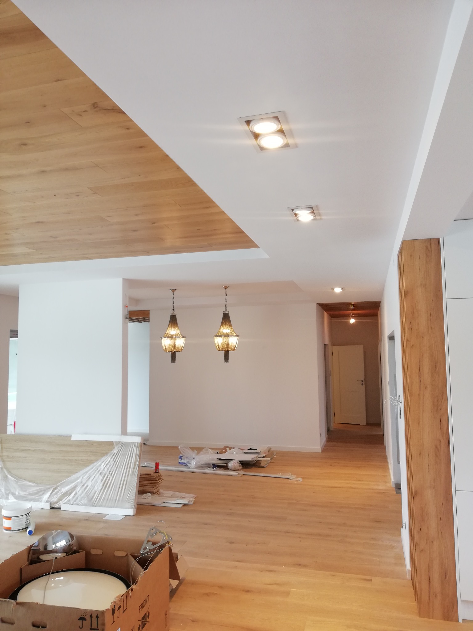 Wnętrze mieszkania w trakcie wykańczania: widoczne zamontowane oświetlenie sufitowe, w tym dwa ozdobne żyrandole i panele LED, częściowo wykończony sufit z drewnianą okładziną, panele podłogowe...