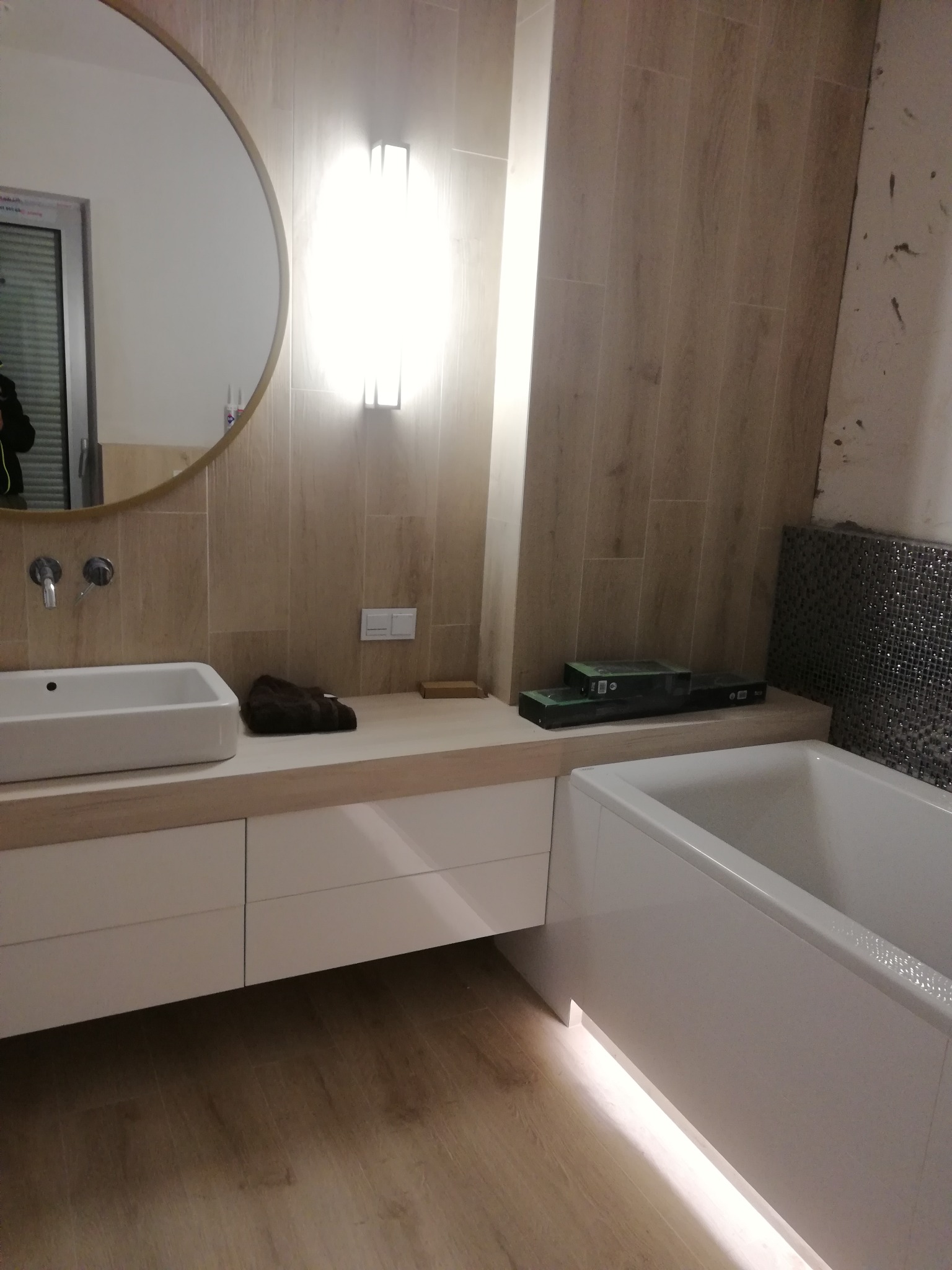 Nowoczesna łazienka z podświetlaną wanną, okrągłym lustrem i drewnopodobnymi płytkami ściennymi. Widoczna biała umywalka nablatowa i oświetlenie punktowe nad lustrem. Część ściany wykończona...