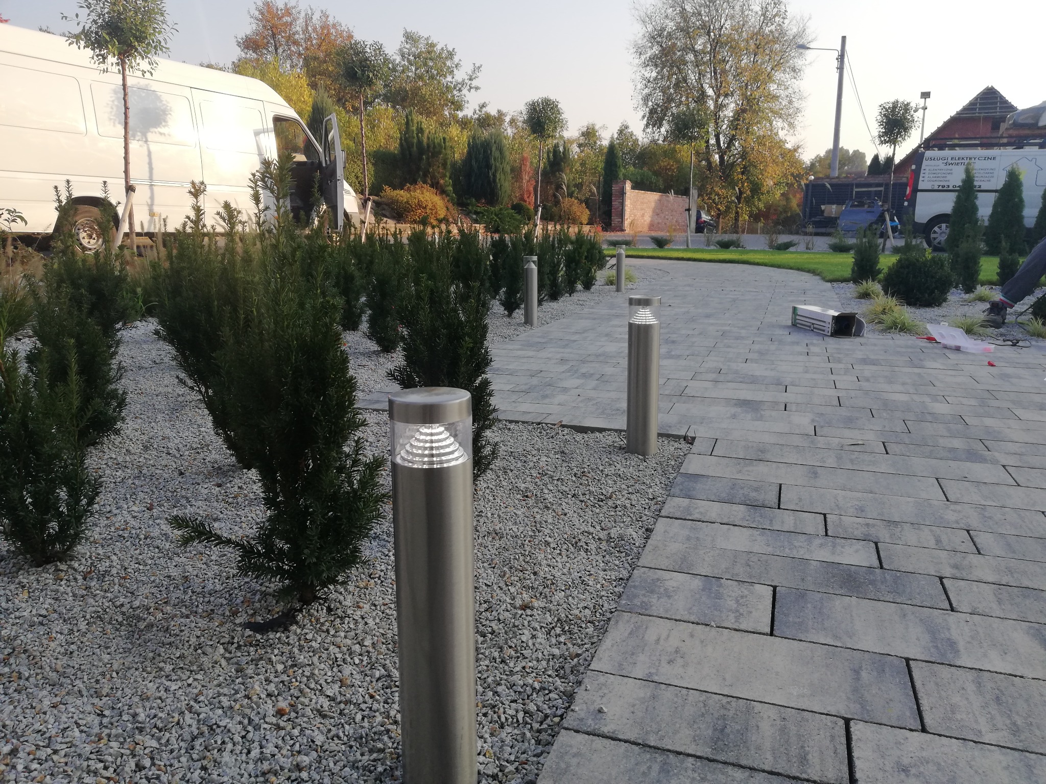 Ścieżka z szarych płyt chodnikowych oświetlona niskimi, metalowymi lampami ogrodowymi, otoczona drobnym białym żwirem i niskimi krzewami, z białym busem w tle.