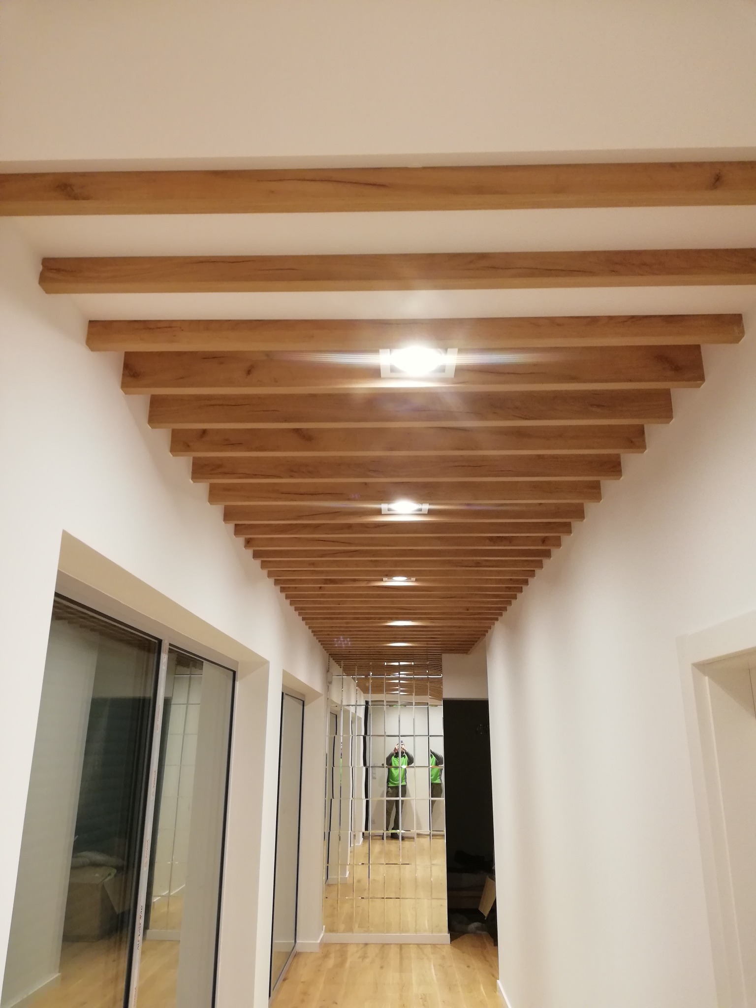 Korytarz z drewnianą podwieszaną konstrukcją sufitową z wbudowanym oświetleniem punktowym, odbicie osoby w lustrzanej ścianie na końcu korytarza.