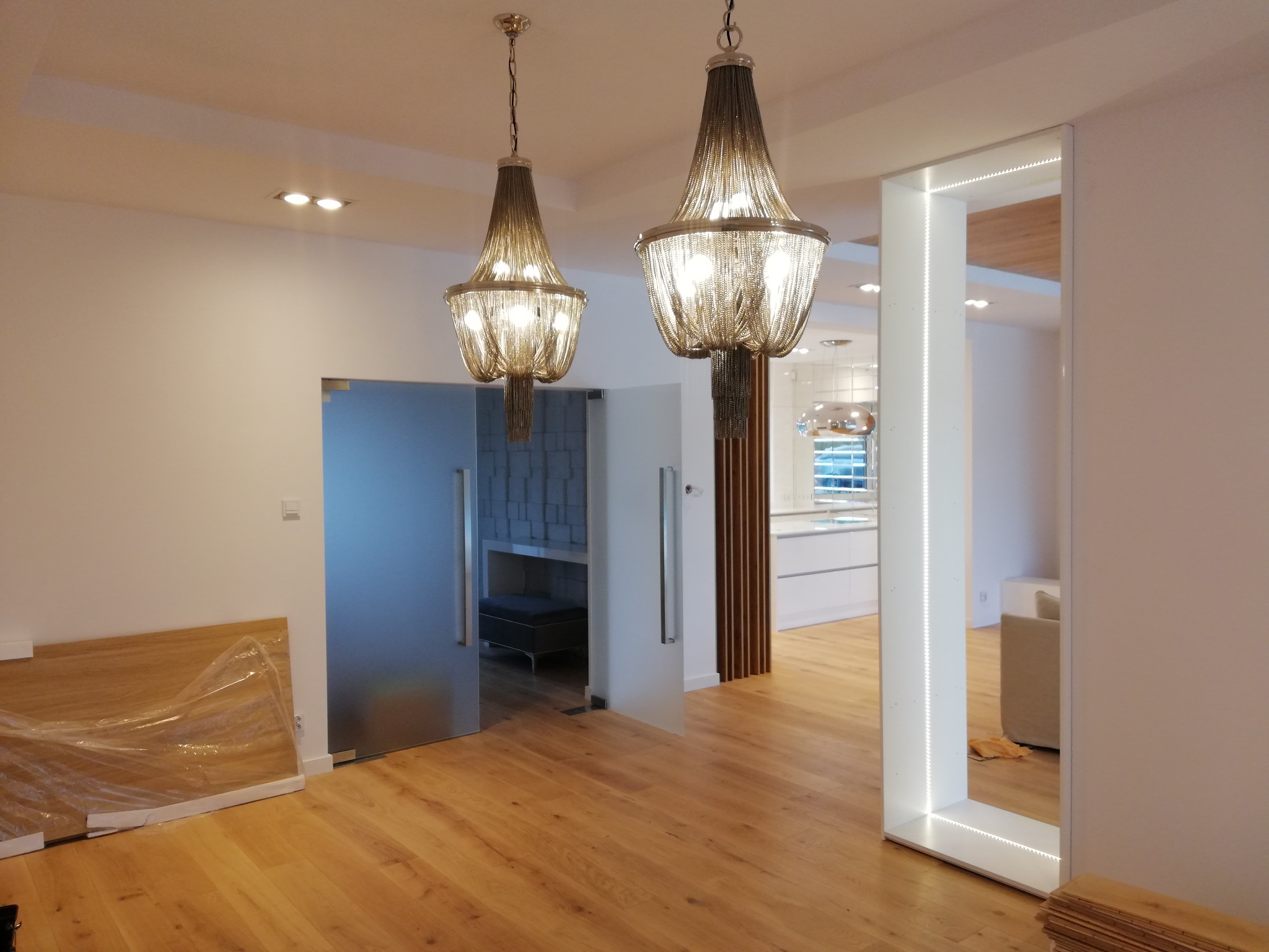Dwa dekoracyjne żyrandole łańcuchowe oświetlające wnętrze domu w Międzychodzie, z podświetlanym LED-ami elementem architektonicznym i parkietem w trakcie układania.