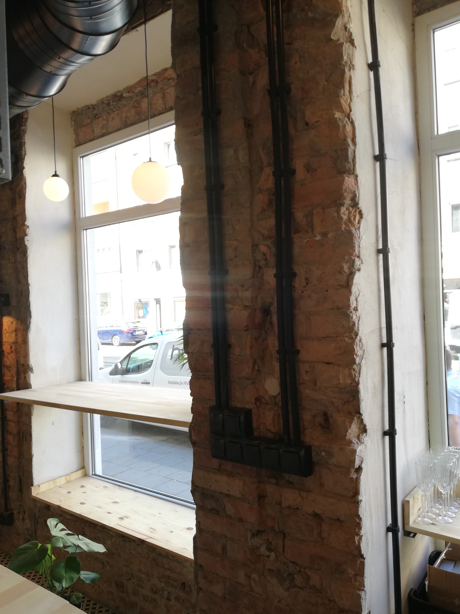 Odsłonięta cegła z czarnymi rurami instalacji elektrycznej i puszkami przy oknie, widoczne lampy wiszące i fragment rośliny doniczkowej.
