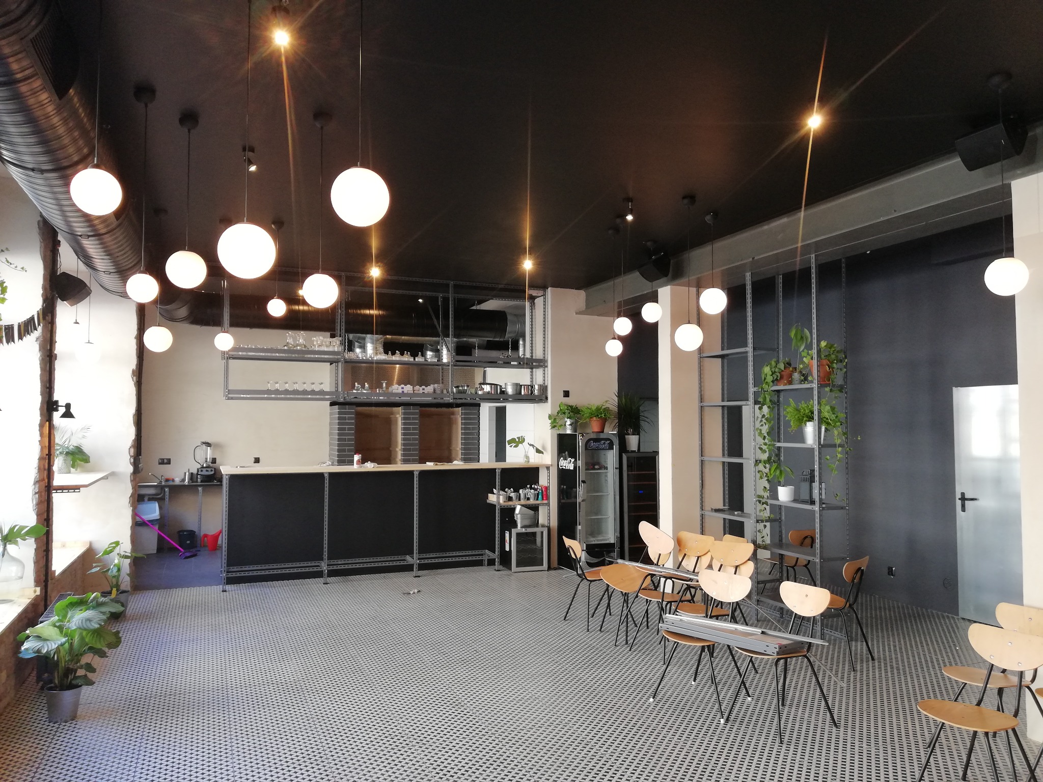 Wnętrze lokalu gastronomicznego z czarnym sufitem, okrągłymi lampami, metalową konstrukcją baru i krzesłami z drewnianymi oparciami, widoczne również rośliny doniczkowe.