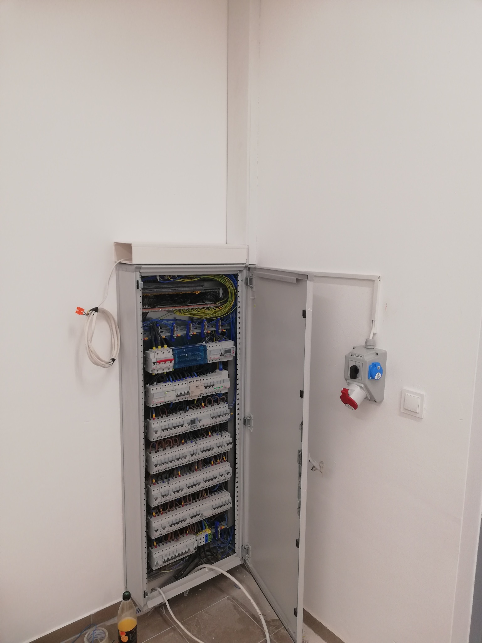 Otwarta szafa elektryczna z bezpiecznikami i okablowaniem, zamontowana w rogu pomieszczenia z białą ścianą, widoczny przewód z końcówkami oraz gniazdo siłowe na ścianie.