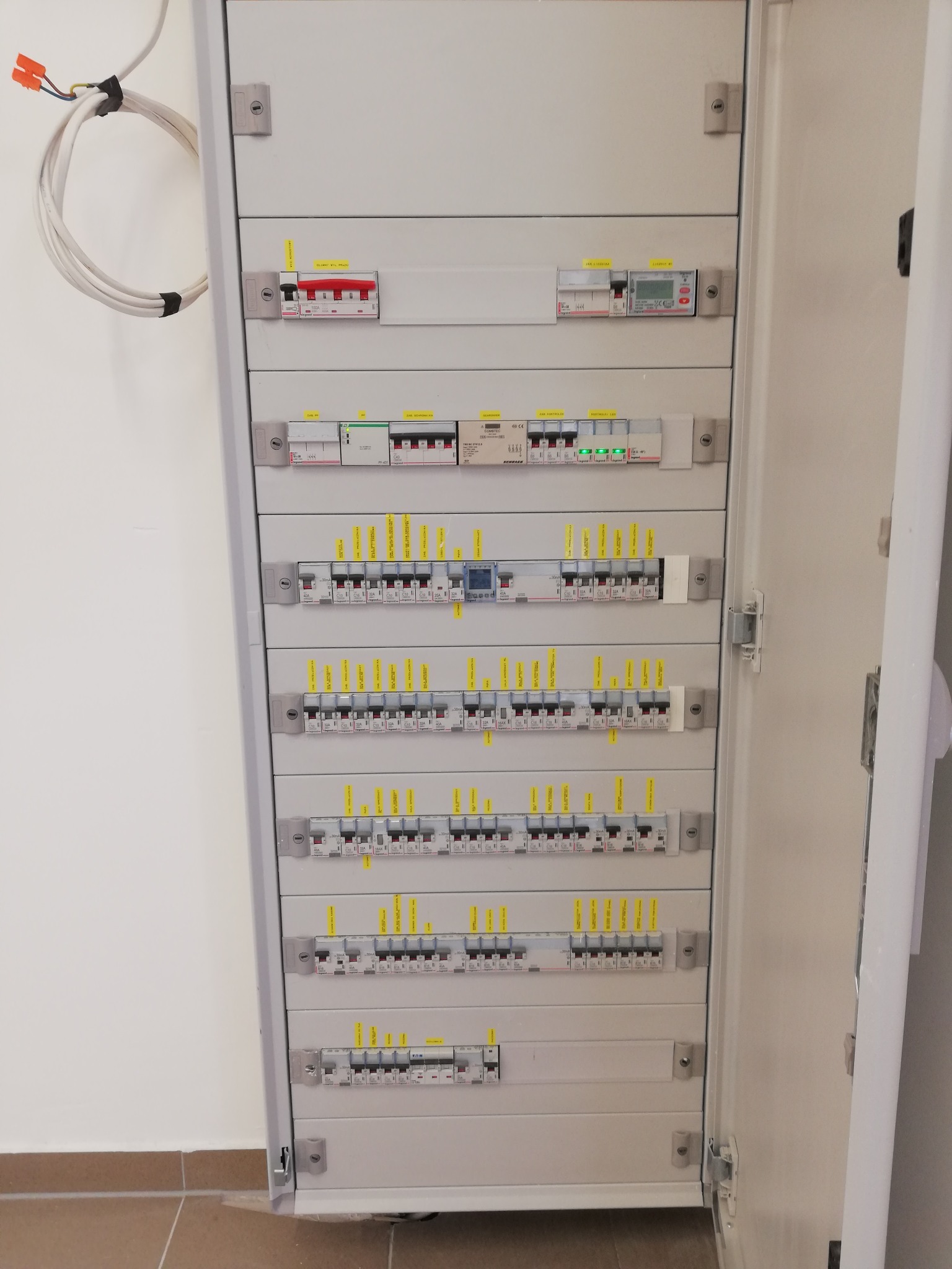 Otwarta szafa rozdzielcza z wieloma zabezpieczeniami nadprądowymi i różnicowoprądowymi, oznaczona żółtymi etykietami, widoczne przewody zasilające.