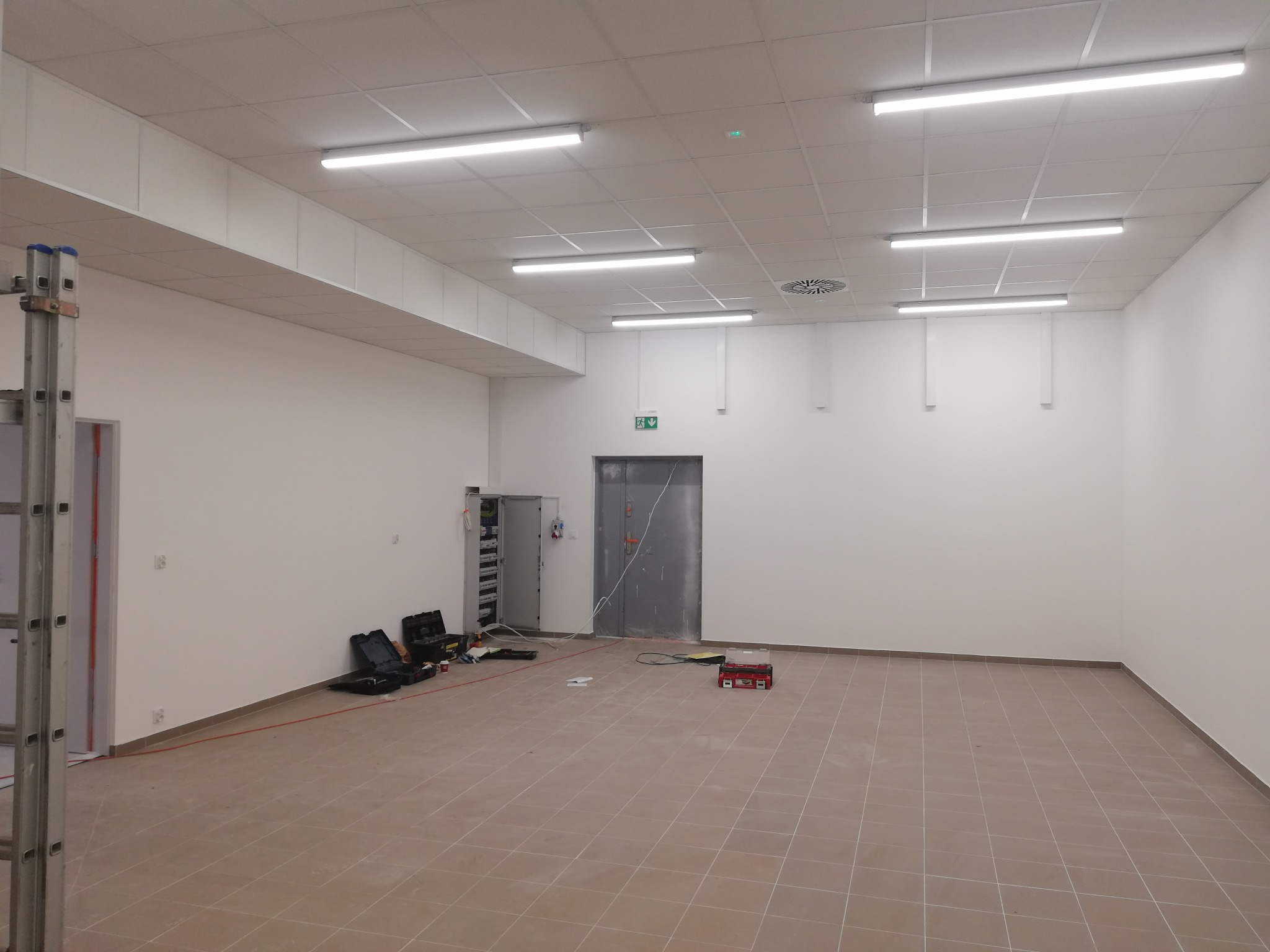 Pusta hala z zamontowanym oświetleniem rastrowym, rozłożone narzędzia i skrzynki narzędziowe, drabina aluminiowa i szafa elektryczna w tle. Widoczny przewód zasilający podłączony do szafy...