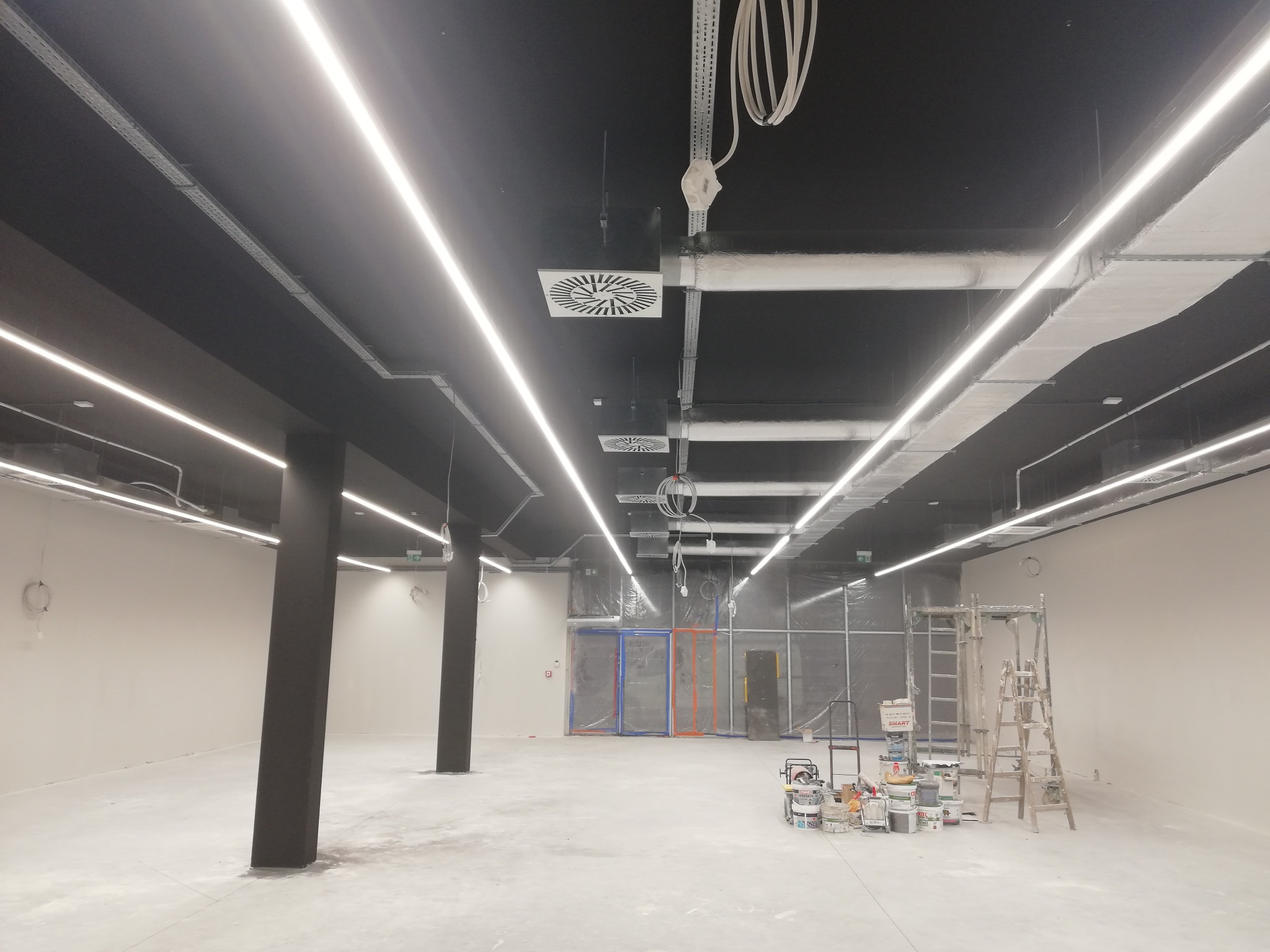 Wnętrze komercyjne w trakcie wykańczania: widoczne podwieszane sufity z oświetleniem liniowym LED, instalacje wentylacyjne, czarne kolumny, drabiny malarskie i puszki z farbą.