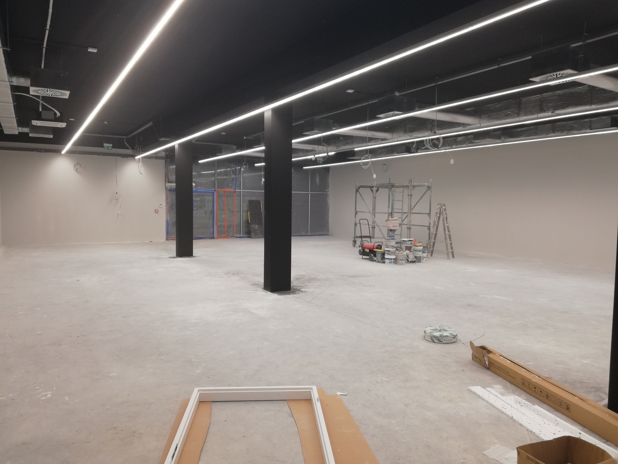 Przestronne wnętrze w trakcie wykończenia, widoczne czarne kolumny, sufit z liniowym oświetleniem LED, rusztowanie, puszki farb i przewody elektryczne na podłodze, ramy okienne w dolnym rogu.