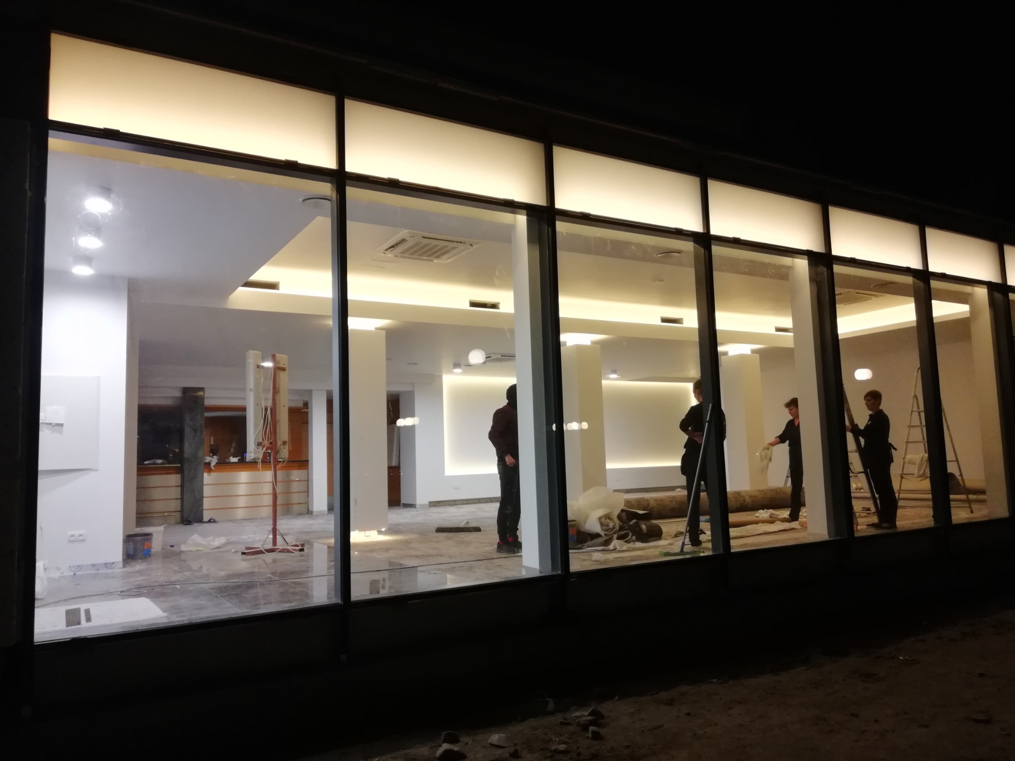 Wnętrze lokalu w trakcie prac wykończeniowych, widoczne przez duże okna. Ekipa montuje oświetlenie sufitowe, w tle rozłożone kable i narzędzia.