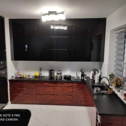 Meble ,,LIZAK,, Dariusz Lizak - Nowoczesna kuchnia z ciemnymi, lakierowanymi szafkami górnymi i dolnymi szafkami z drewna egzotycznego, czarny blat, marmurowa podłoga, widoczne akcesoria kuchenne.